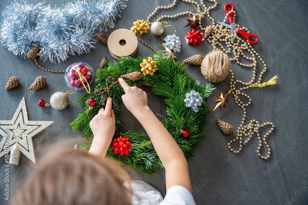 Fototapeta premium Little girl decorating a Christmas wreath while sitting at the table, top view. Capturing a hands-on festive activity from an overhead perspective at home.