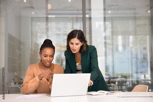 Two multiethnic female coworkers working together at laptop, pointing at screen, discussing task. Young Black professional asking colleague for advice, feedback, showing content on computer