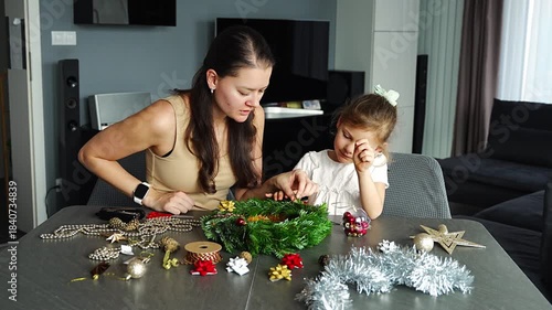 Mother and her little daughter decorating a Christmas wreath together at home. Strengthening family bonds through shared festive traditions and creative seasonal activities.