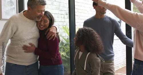 Diverse family arriving at glass pivot door exchanging hugs, handing green gift, patting son's head