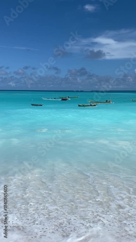 Vertical footage of crystal-clear turquoise ocean water on a white sandy beach with several small wooden fishing boats anchored offshore. Tropical travel concept.