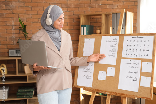 Young Muslim woman with laptop learning Arabic language online near pin board...