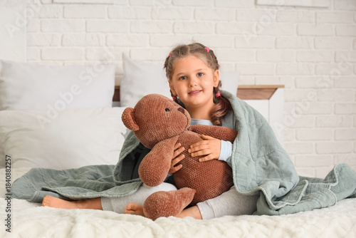Cute little happy girl in pajamas with teddy bear and plaid sitting on bed at...