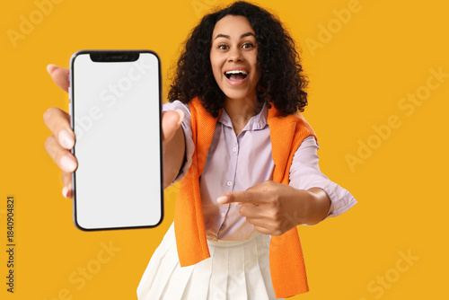 Stylish young African-American woman pointing at blank mobile phone on yellow...