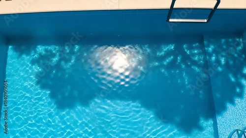 Swimming pool with shimmering blue water, sunlight reflections, and tree shadows creating a serene summer scene.
