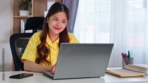 Asian Young woman smiling and focused working on laptop at home desk with notebook, pen and smartphone in warm natural light for remote work office style setup