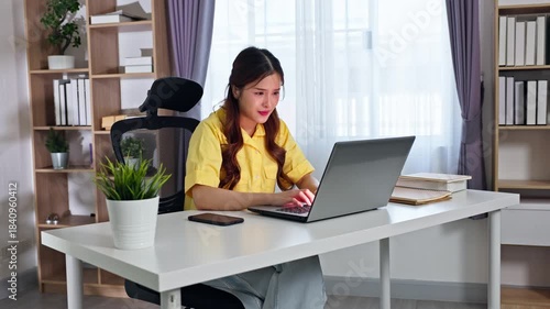 Asian Smiling woman yellow shirt video chatting laptop bright home office desk with plant and books, young woman working laptop tidy home office desk with plant smartphone notebook and bookshelf