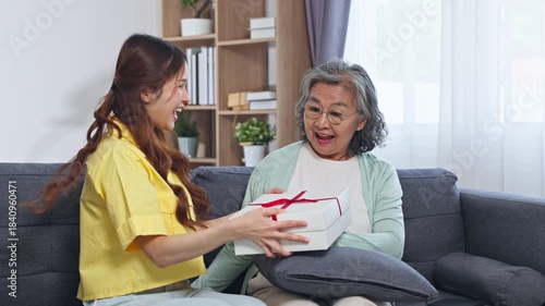 Asian grandmother and granddaughter embrace sofa cozy living room as young woman gives wrapped gift present red ribbon hugging and laughing while holding cushion sharing joyful family hug and surprise