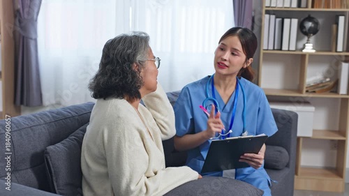 Asian Nurse consulting elderly woman living room couch with clipboard and stethoscope reviewing medical notes, compassionate home care consultation showing concern and support during medical