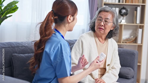 Asian Elderly woman caregiver consultation about medication on living room sofa compassionate attentive support nurse showing medication explaining dosage calm home care concerned expression
