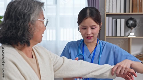 Asian nurse caregiver home visit medical assessment examining elderly woman arm with gentle compassionate examination and supportive care during home medical visit