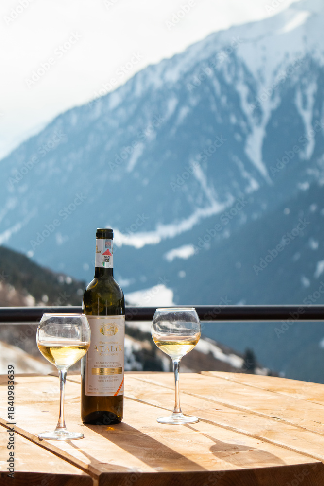 Fototapeta premium Osh, Kyrgyzstan - December 13, 2024: A bottle of Atalyk white wine and two glasses stand on a wooden table with a dramatic, snowy, mountainous background
