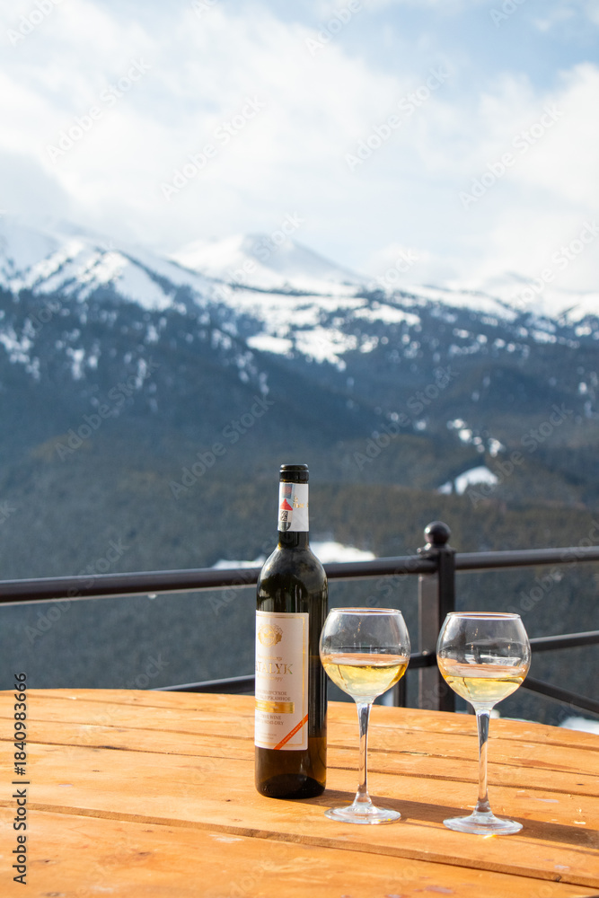 Fototapeta premium Osh, Kyrgyzstan - December 13, 2024: A bottle of Atalyk white wine and two glasses stand on a wooden table on a terrace overlooking vast, snow-covered mountains