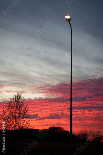 silhouetted streetlight stands tall against a dramatic sunset sky with vibrant red and orange hues, contrasting with silhouetted trees and a peaceful, serene evening landscape, idyllic, majestic