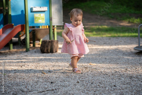 toddler in a pink dress explores playground on sunny day, clutching small sticks, surrounded by play equipment and textured gravel ground with greenery in background, joyful, texture, environment