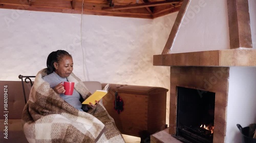 Senior Black woman drinking hot coffee and reading a book by the fireplace at home