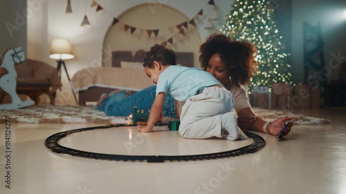Merry family weekend in Christmas holidays, african american mother and son. Happy little boy and his mom sitting on floor in cozy bedroom, playing toy railway, celebrating Xmas and new year together