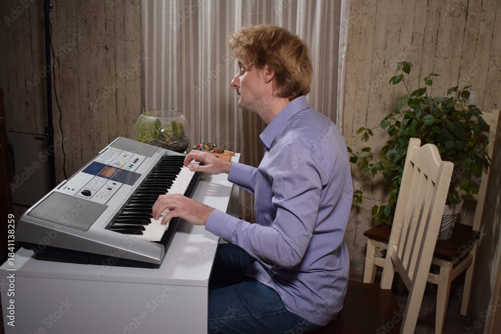 Obraz premium A young white man plays an electronic synthesizer indoors, immersed in his creative hobby. Focused on shaping tones and melodies, he explores musical expression and artistry in a calm home setting. 