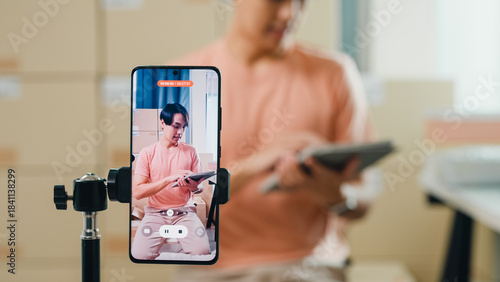 фотографія Close up youth Asia male owner small business online shop sit front of phone look at camera live streaming see comment on digital tablet from customer sales promotion at retail online shop at home
