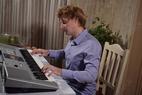 A young white man plays an electronic synthesizer indoors, immersed in his creative hobby. Focused on shaping tones and melodies, he explores musical expression and artistry in a calm home setting.
