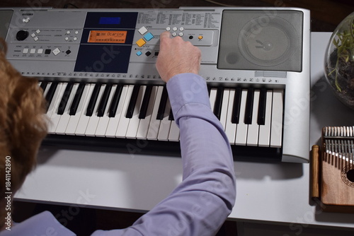 A young white man plays an electronic synthesizer indoors, immersed in his creative hobby. Focused on shaping tones and melodies, he explores musical expression and artistry in a calm home setting.
