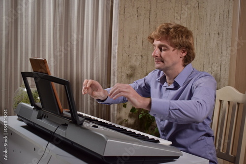 A young white man plays an electronic synthesizer indoors, immersed in his creative hobby. Focused on shaping tones and melodies, he explores musical expression and artistry in a calm home setting.

