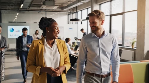 Two colleagues stroll and chat in a bright, modern office as teams collaborate nearby. and teamwork