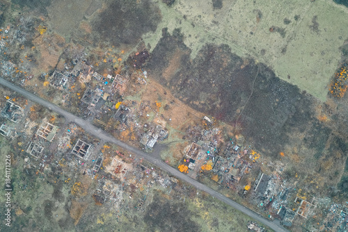 Destroyed settlement ruins and scattered rubble along the central road dividing forest and field. War zone urban destruction pattern - Aerial drone shot