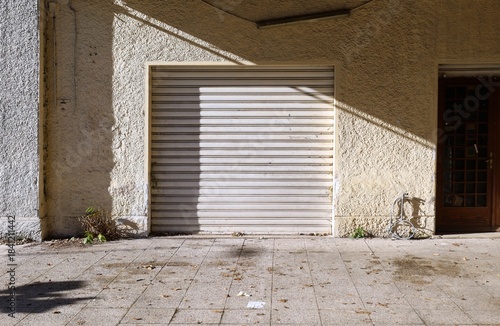 Long time closed store with white rolling shutters down and door on the right. Dirty tiled pavement. Background for copy space