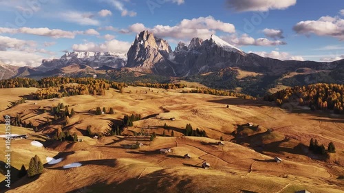 Aerial view of Alpe di Siusi (Seiser Alm) and the Sassolungo group in the Dolomites. Majestic alpine peaks covered in light snow glow in warm evening light above autumn forest and green meadows.