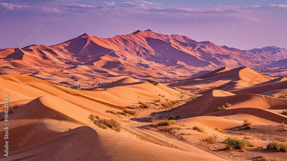 Naklejka premium Vibrant orange sand dunes under a purple sky landscape desert terrain with scattered vegetation and winding tracks leading through arid wilderness during sunset scenery