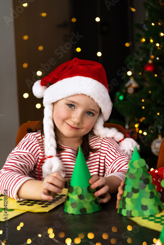 Happy girl smiles and make diy Christmas tree from green paper. Child creates holiday decorations for Christmas. Craft for New Year