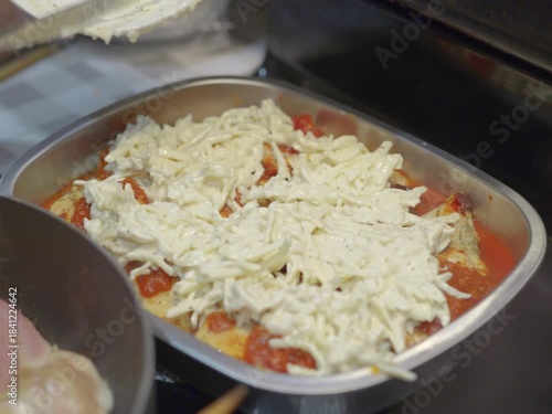 In a close-up shot, a black womans hand is seen spreading shredded cheese over a tomato-sauced dish in a metal baking tray, preparing it for baking.