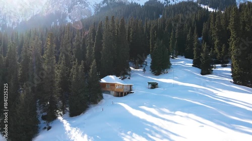Aerial view of a secluded wooden cabin surrounded by pine trees in a snowy mountain landscape with a strong red sun flare