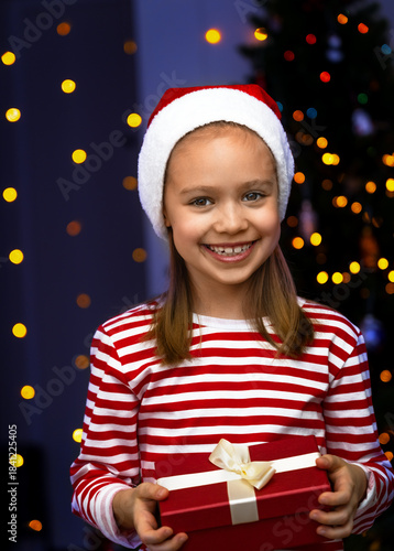 Portrait of happy girl in santa hat with gift box looks at camera and smiles on blue background of Christmas tree. Child with New Years present
