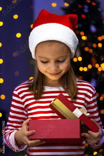 Happy little girl in santa hat opens gift box with light next to Christmas tree. Child with New Years present.