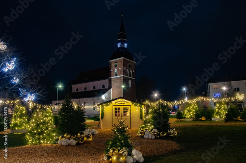Valmiera,Latvia-December 04.2025-Festive Christmas lights, decorated trees, and a small holiday house glow warmly in front of a historic church on a winter night.