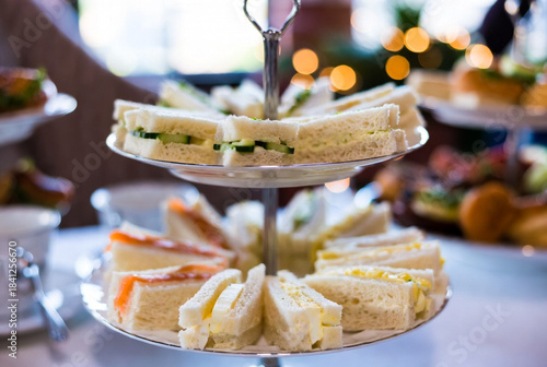 A two-tier serving tray presents an assortment of finger sandwiches—cucumber, egg salad, salmon, and cheese—set for a festive buffet. Warm bokeh lights create a cozy, inviting atmosphere.