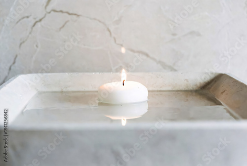 Single white candle floats in shallow water-filled marble tray, casting soft glow. Serene scene evokes relaxation, meditation, peaceful ambiance suitable for spa, wellness, spiritual imagery.