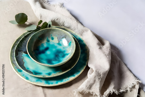 Serene flat lay of turquoise glazed ceramic plates and bowls stacked on natural linen, accented by eucalyptus. Elegant tableware scene evokes calm dining, handmade craftsmanship, earthy, modern home
