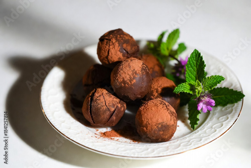 Rich, cocoa-dusted chocolate truffles sit on a white plate, accented by fresh mint and a delicate purple flower. A gourmet, indulgent treat suited for dessert, gifting, or festive gatherings.