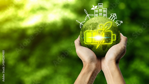 Hands holding a glowing green globe with a battery and leaf icon, surrounded by outlines of wind turbines and eco buildings, representing bio energy and sustainable living. Concept of Bio Energy