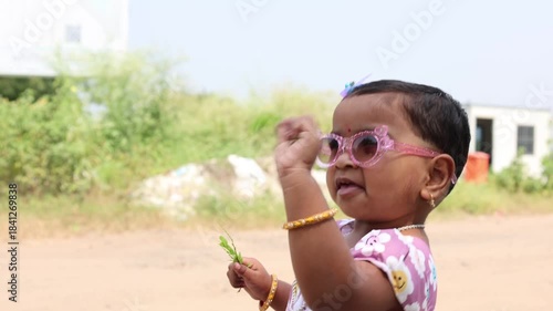 An Indian girl child wearing pink sunglasses and a floral dress holding a small plant outdoors, smiling gently in bright daylight with soft greenery and buildings creating a playful natural background