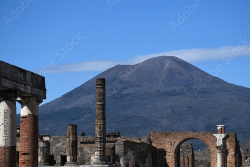 Mt Vesuvius rises up above the ruins of the Forum in Pompeii. A road can be seen zig-zagging across the mountain. The sky is blue.