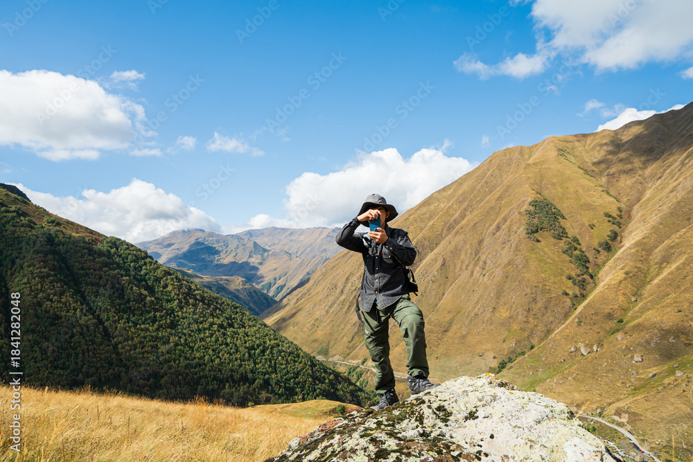 Fototapeta premium Successful hiker taking a photo on a rocky peak in Juta Village, Georgia. Mountain climbing adventure with panoramic views, sunny blue sky, and active outdoor lifestyle in the Caucasus Mountains.