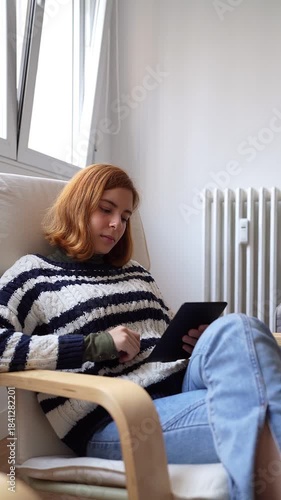 Relaxed teenage girl sitting in a comfortable armchair reading an e-book by the window at home