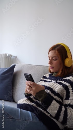 Relaxed young woman with yellow headphones using a mobile phone while sitting on a comfortable sofa