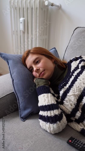 Sad redhead woman with a pensive expression lying on the sofa at home, feeling bored and lonely