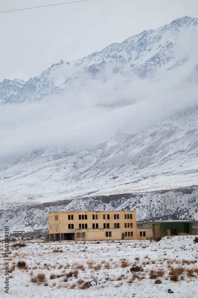 Fototapeta premium A derelict multi-story building standing in a snowy field with low clouds covering the massive mountain range behind it