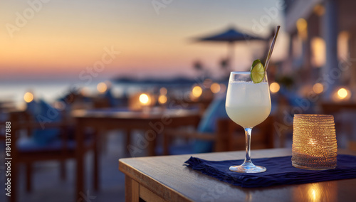 Sophisticated pale yellow cocktail in an elegant stemmed glass garnished with a lime slice and metal straw on a wooden table with a navy cloth and softly glowing candle at sunset